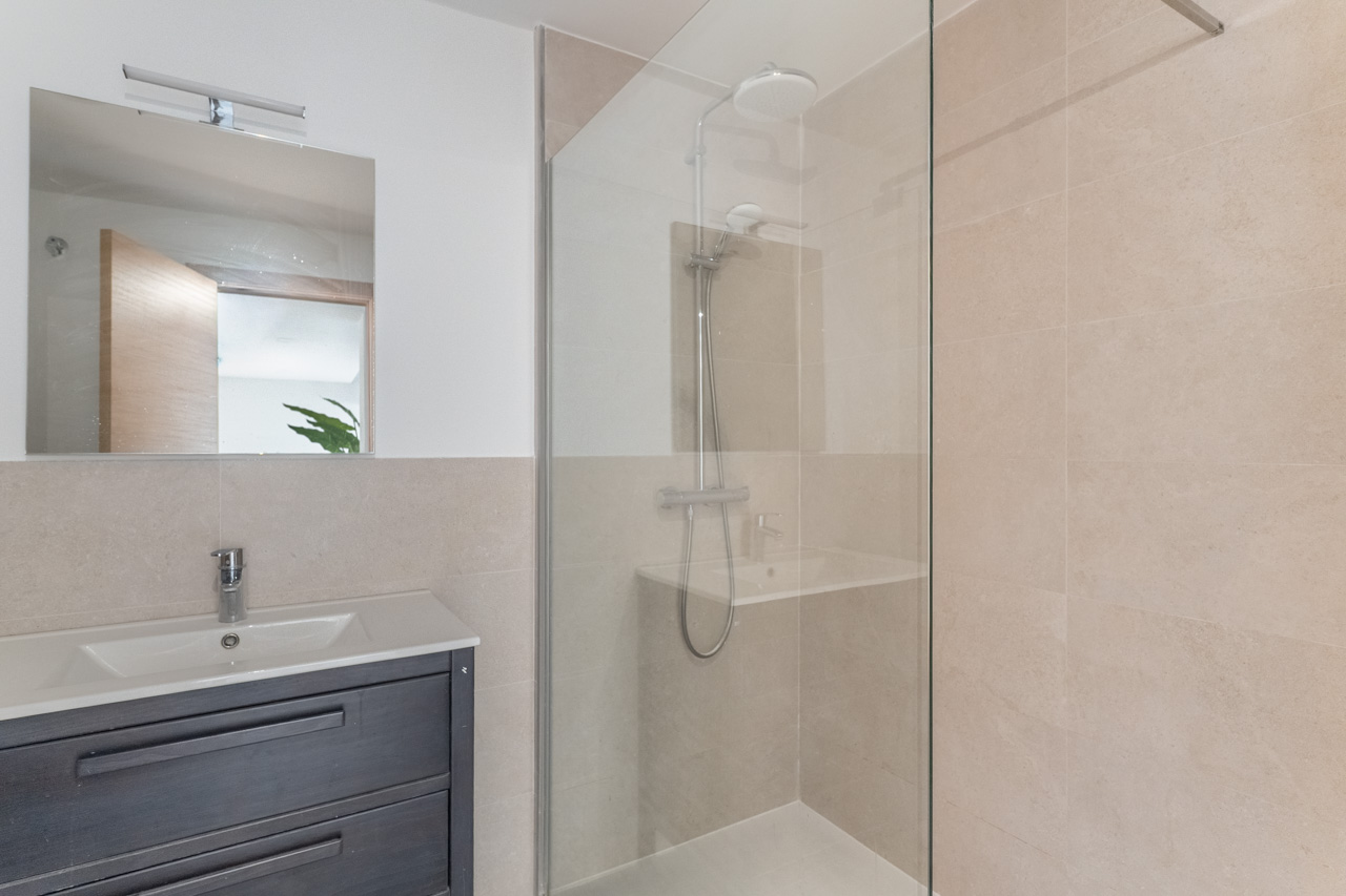 Salle de bain moderne avec douche à l'italienne vitrée, carrelage mural beige et meuble vasque en bois sombre avec miroir carré.