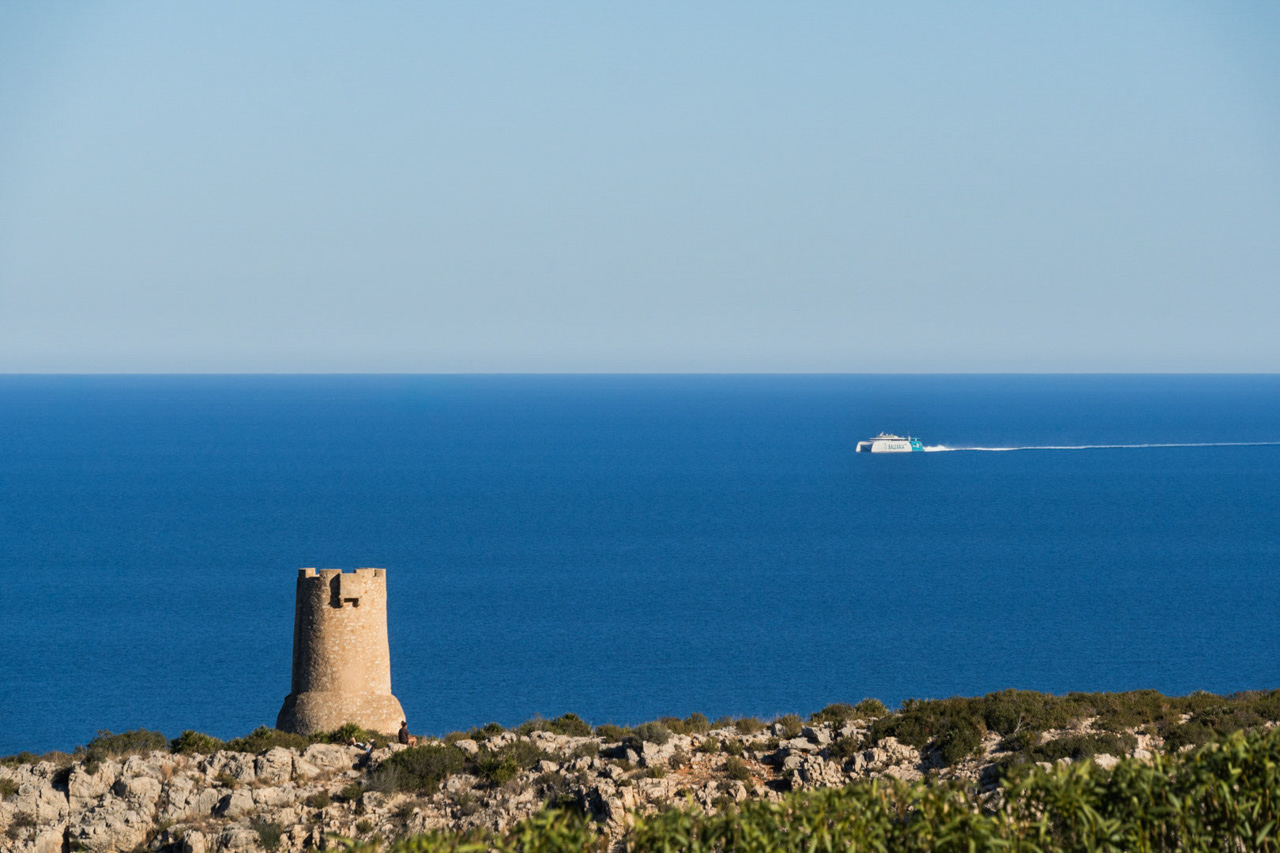 Vue d'une tour de guet historique en pierre sur une falaise rocheuse surplombant la mer Méditerranée, avec un ferry traversant l'horizon par temps clair.