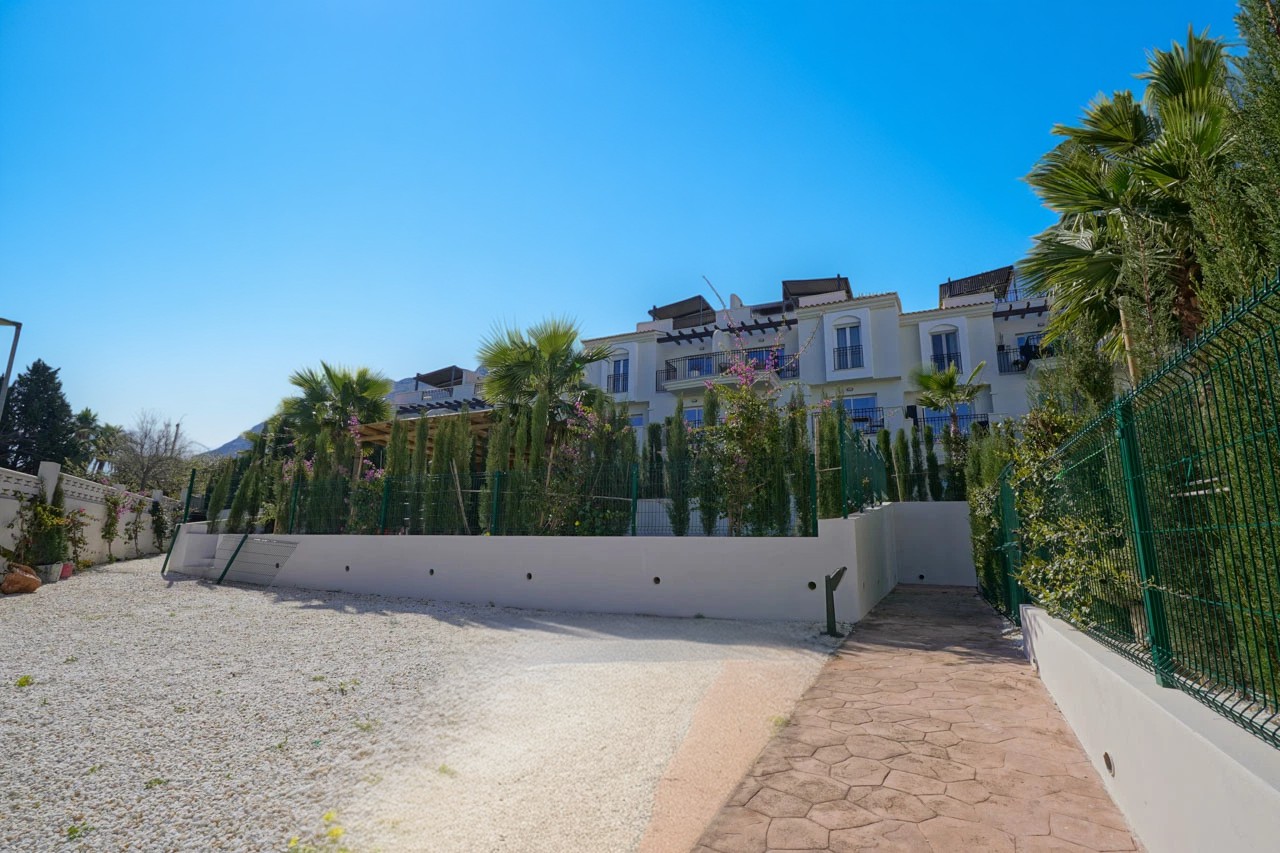 Vue extérieure d'un complexe résidentiel blanc avec balcons privés, palmiers et un chemin pavé bordant une zone de gravier sous un ensoleillement direct.