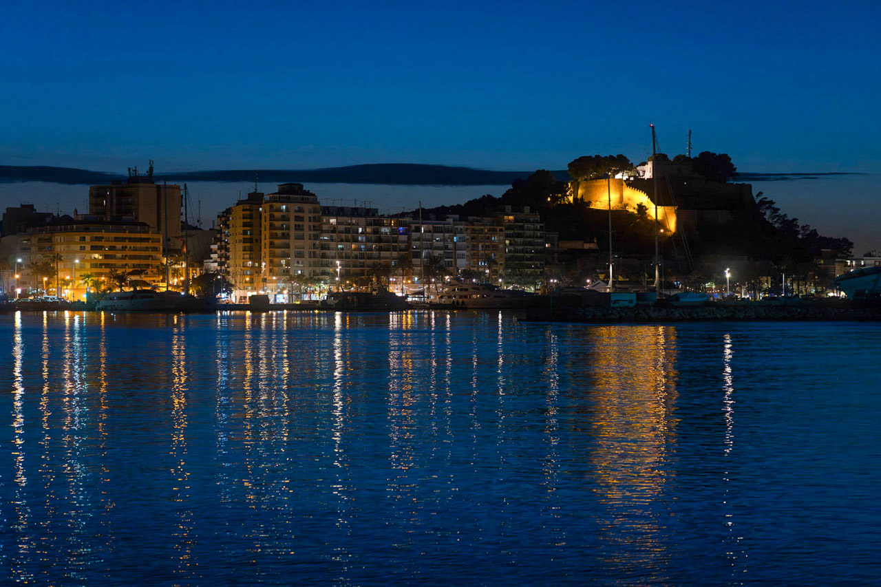 Vue nocturne de la ligne d'horizon côtière et du port, avec le château historique illuminé sur la colline et des reflets dorés sur l'eau calme.