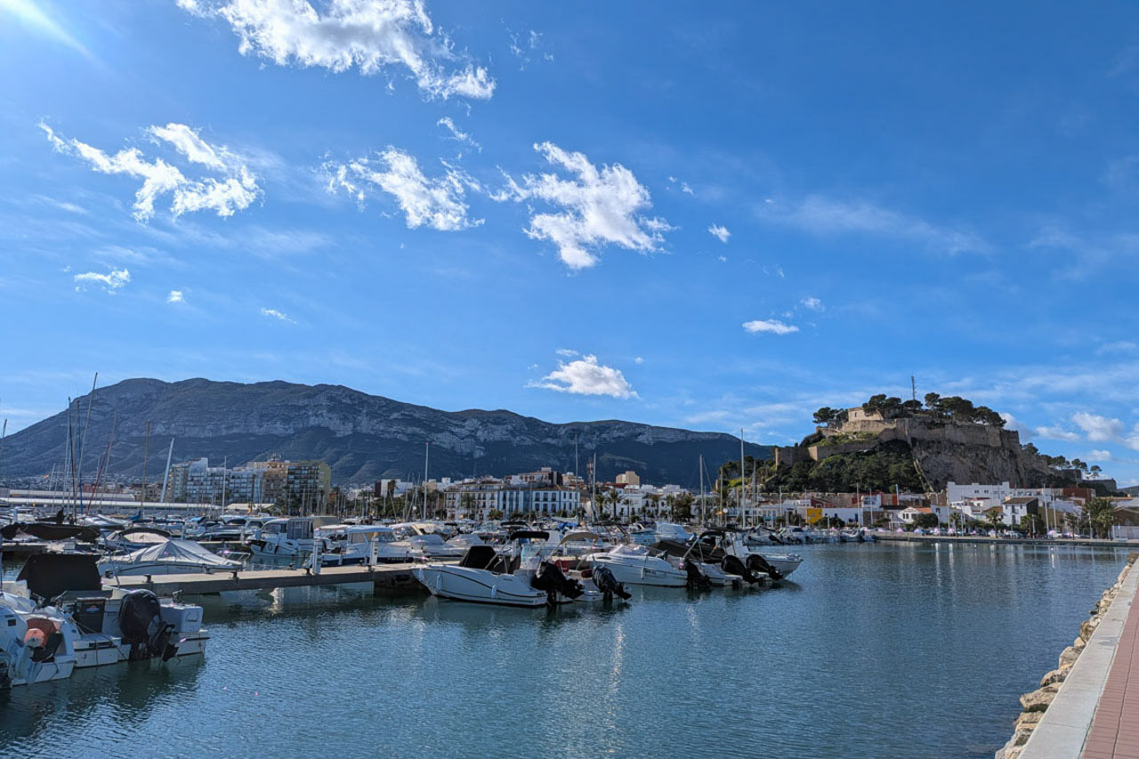 Vue panoramique sur le port de plaisance de Dénia, avec ses bateaux amarrés, le château historique sur la colline et le massif du Montgó en arrière-plan.
