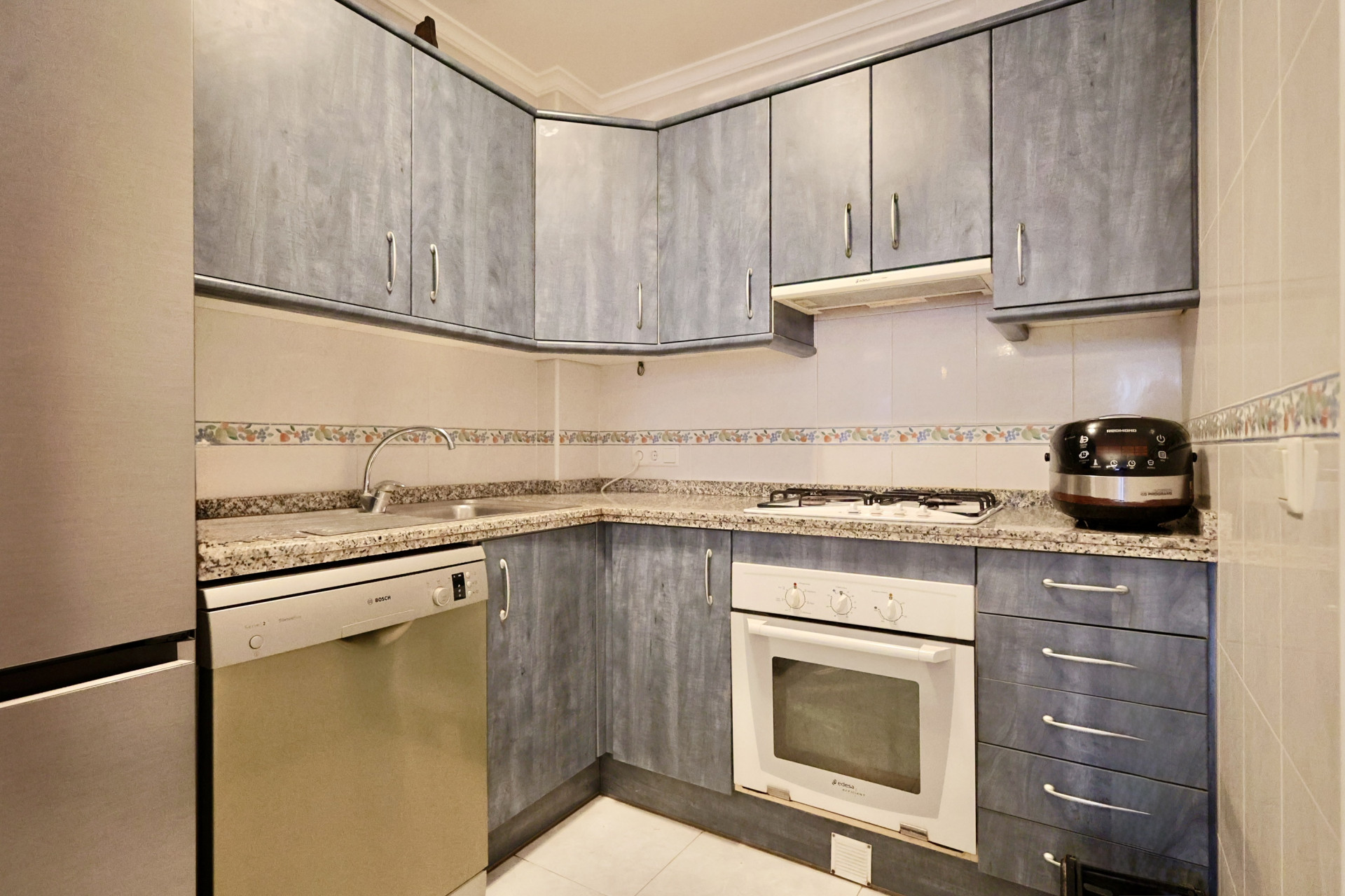 Cuisine équipée avec placards bleus, plan de travail en granit, lave-vaisselle Bosch et four blanc. Crédence en carrelage clair ornée d'une frise décorative florale.