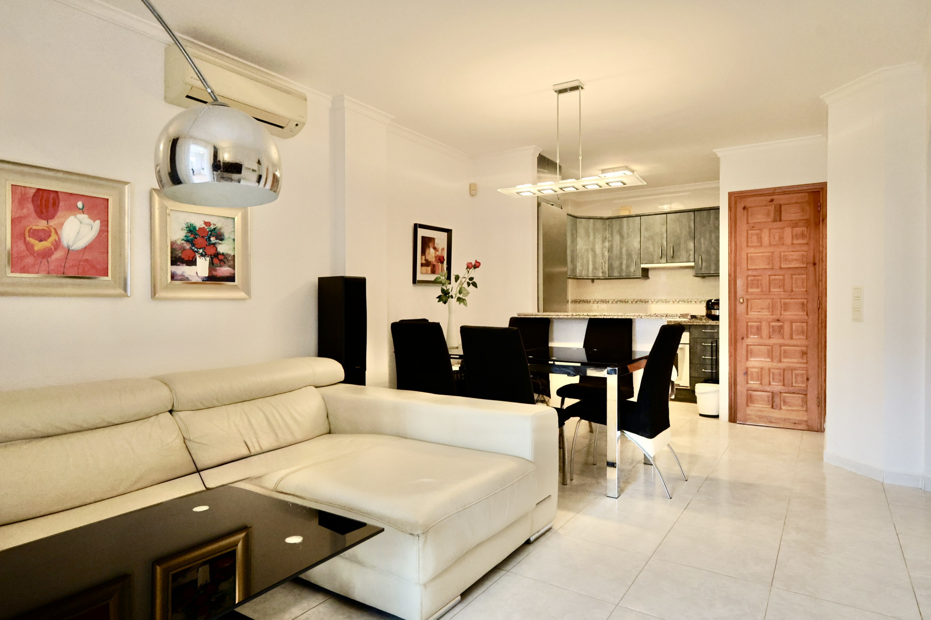 Espace de vie ouvert avec canapé en cuir blanc, table à manger en verre noir et cuisine équipée de meubles gris et plans de travail en granit.