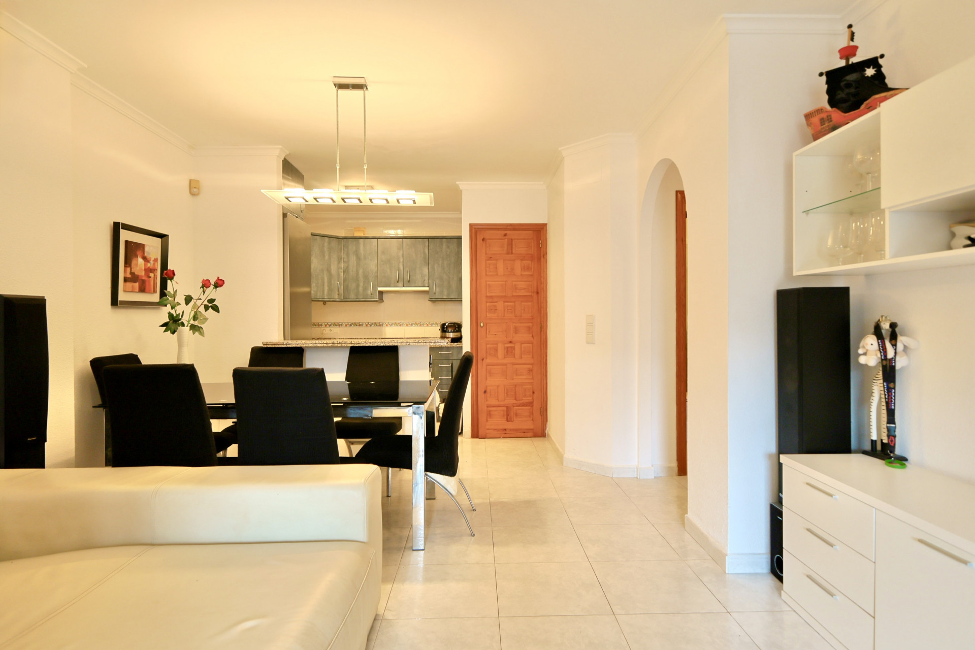 Espace de vie ouvert avec salle à manger aux chaises noires, luminaire moderne et cuisine grise. Carrelage clair, porte en bois et passage voûté.