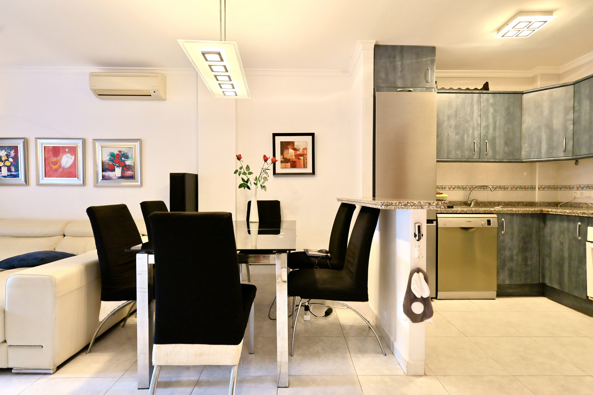 Espace de vie ouvert comprenant un coin repas avec table en verre et chaises noires, jouxtant une cuisine aux meubles bleus et plan de travail en granit.