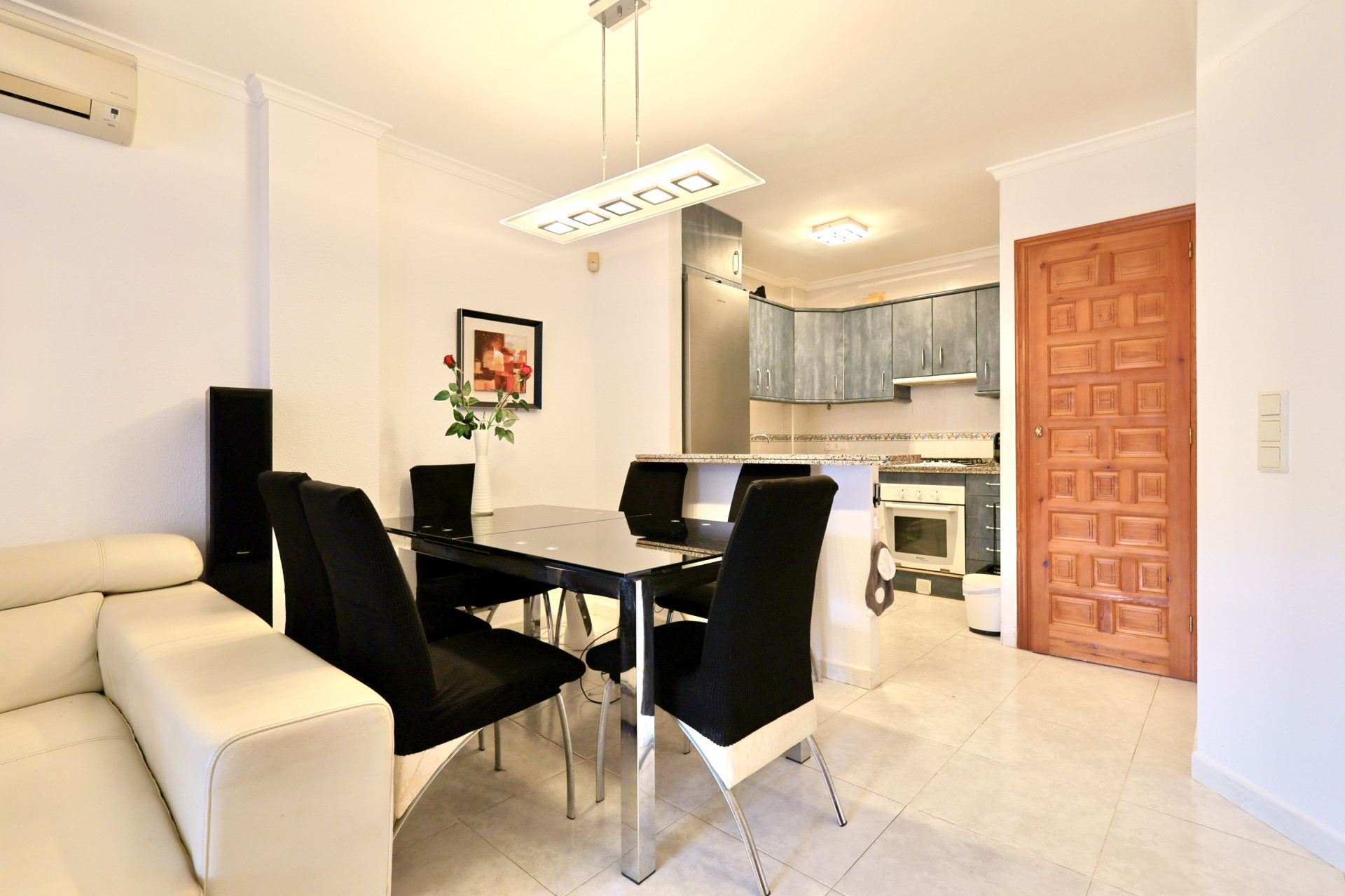 Espace de vie ouvert comprenant une table à manger en verre noir, une cuisine intégrée avec meubles bleus et plans de travail en granit, et un sol en céramique claire.