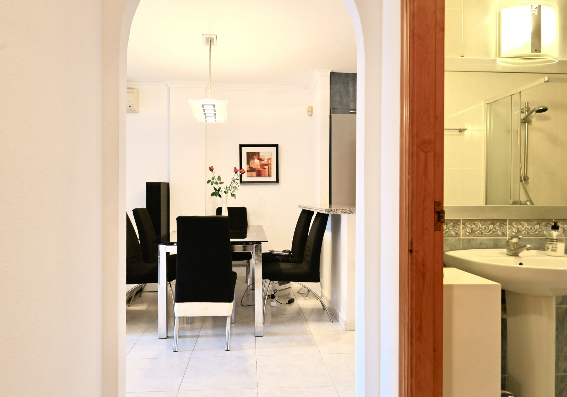 Vue à travers une arche sur la salle à manger avec table en verre et chaises noires, jouxtant une salle d'eau avec lavabo sur colonne et douche.