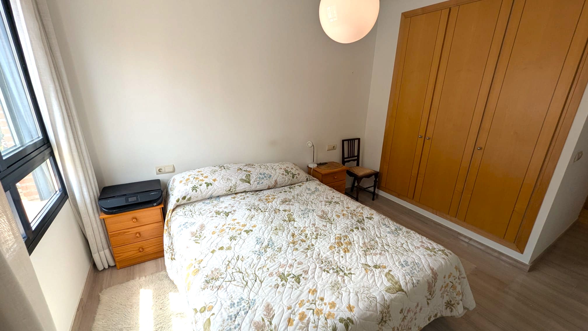 Chambre avec armoire encastrée en bois à quatre portes, lit double et tables de chevet assorties. Sol en parquet clair et fenêtre à cadre noir avec rideaux neutres.