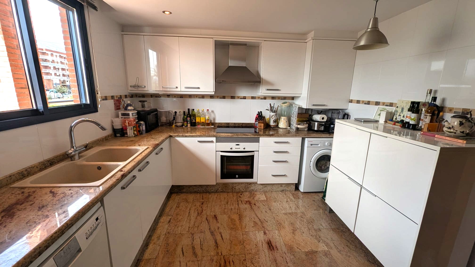 Cuisine fonctionnelle avec placards blancs, plans de travail en granit et sol en pierre naturelle. Comprend des appareils intégrés et une fenêtre à cadre sombre.