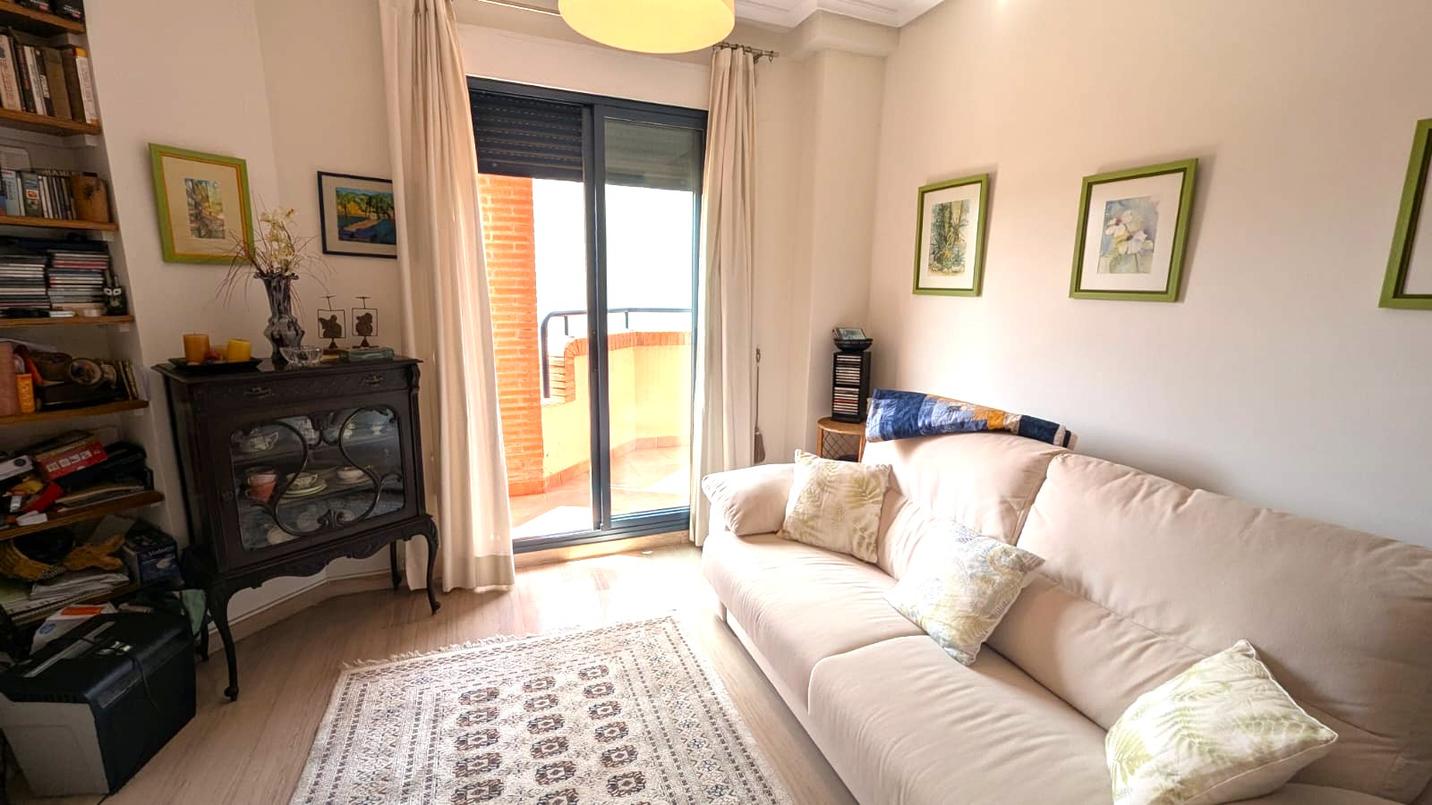 Salon lumineux avec canapé beige, vitrine ancienne en bois sombre et accès direct à un balcon par une baie vitrée coulissante.