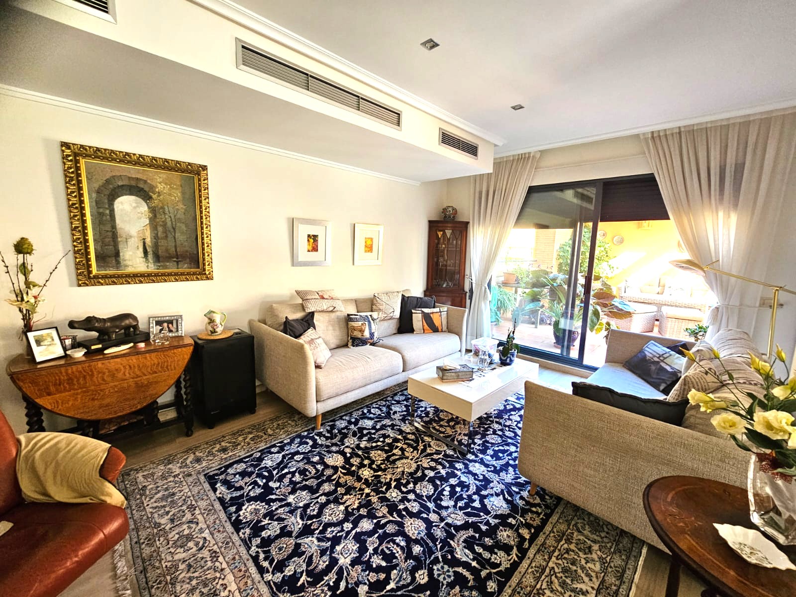 Salon spacieux avec canapés en tissu, tapis ornemental bleu et accès direct à la terrasse. Équipé de climatisation par conduits et d'un éclairage encastré au plafond.