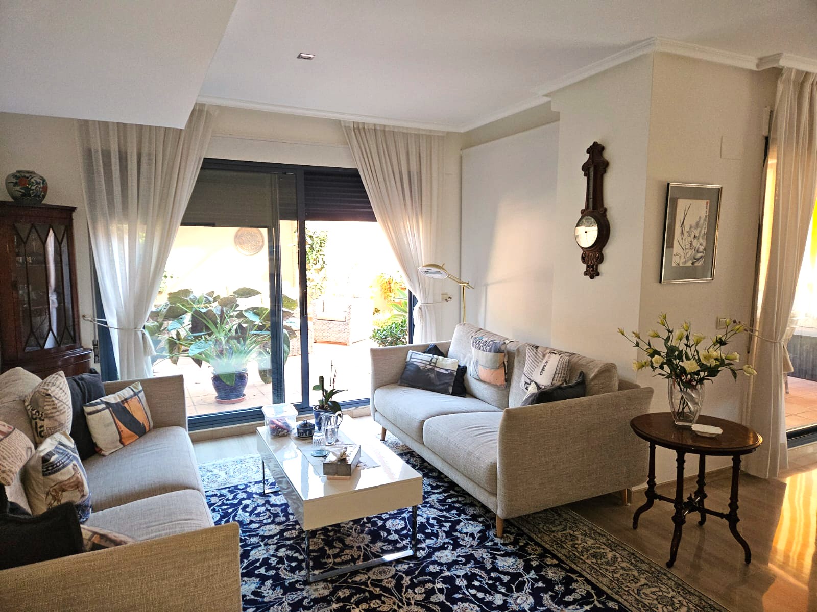 Séjour lumineux avec deux canapés neutres, un tapis bleu à motifs et de grandes baies vitrées coulissantes ouvrant sur une terrasse privée verdoyante.