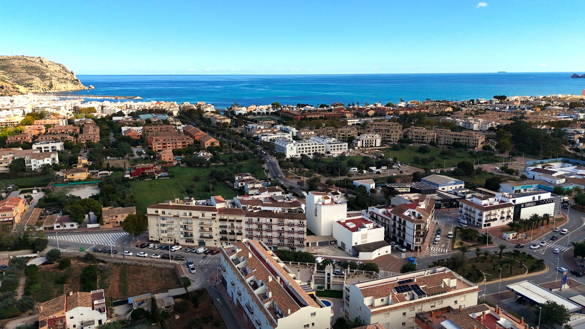 Vue panoramique surélevée du paysage urbain de Jávea, mettant en valeur l'architecture méditerranéenne, la zone portuaire et la mer bleue sous un ciel dégagé.