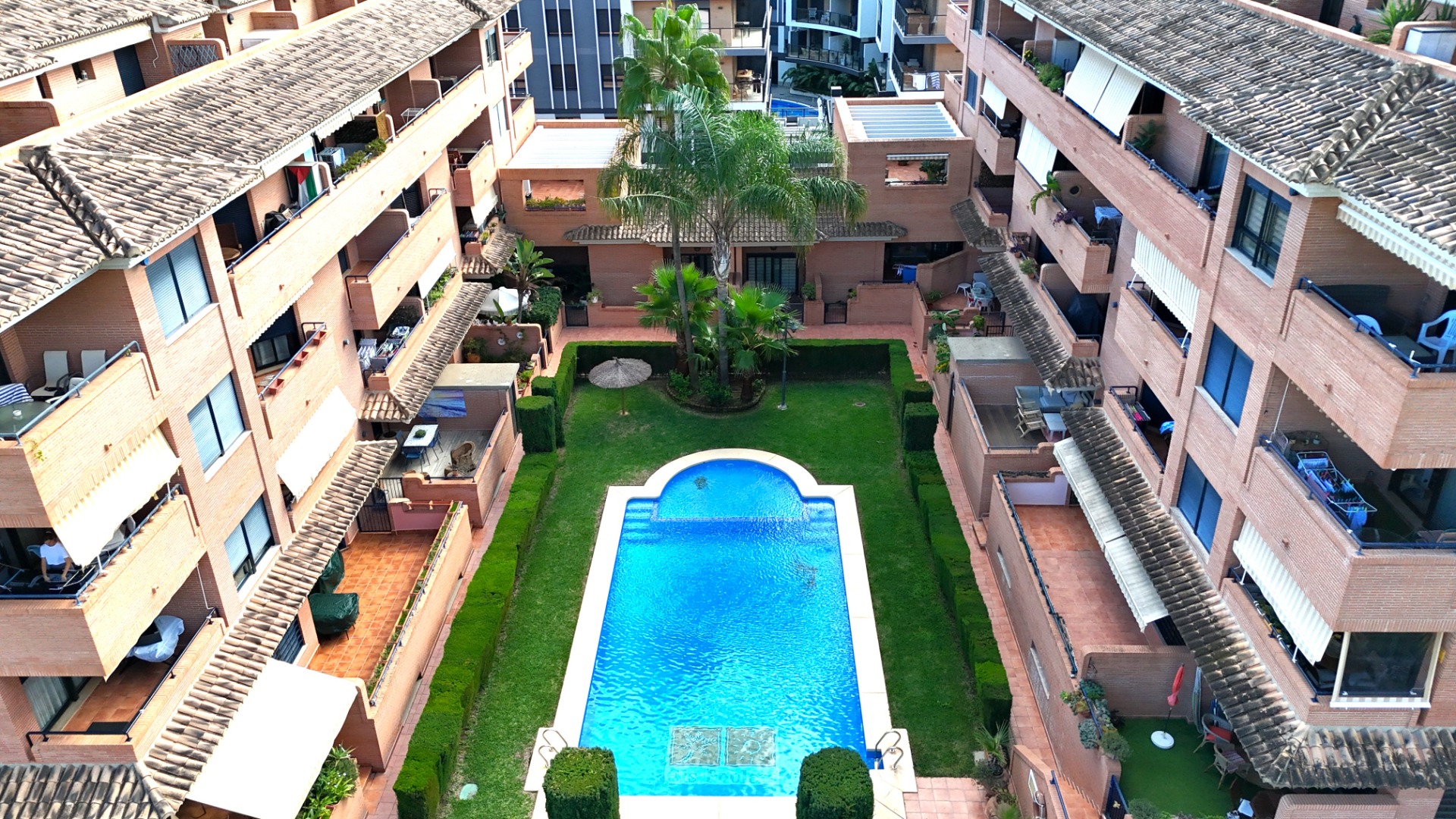 Vue surélevée de l'espace commun avec piscine, pelouse soignée et palmiers, entouré d'immeubles en briques dotés de terrasses privées à Jávea.