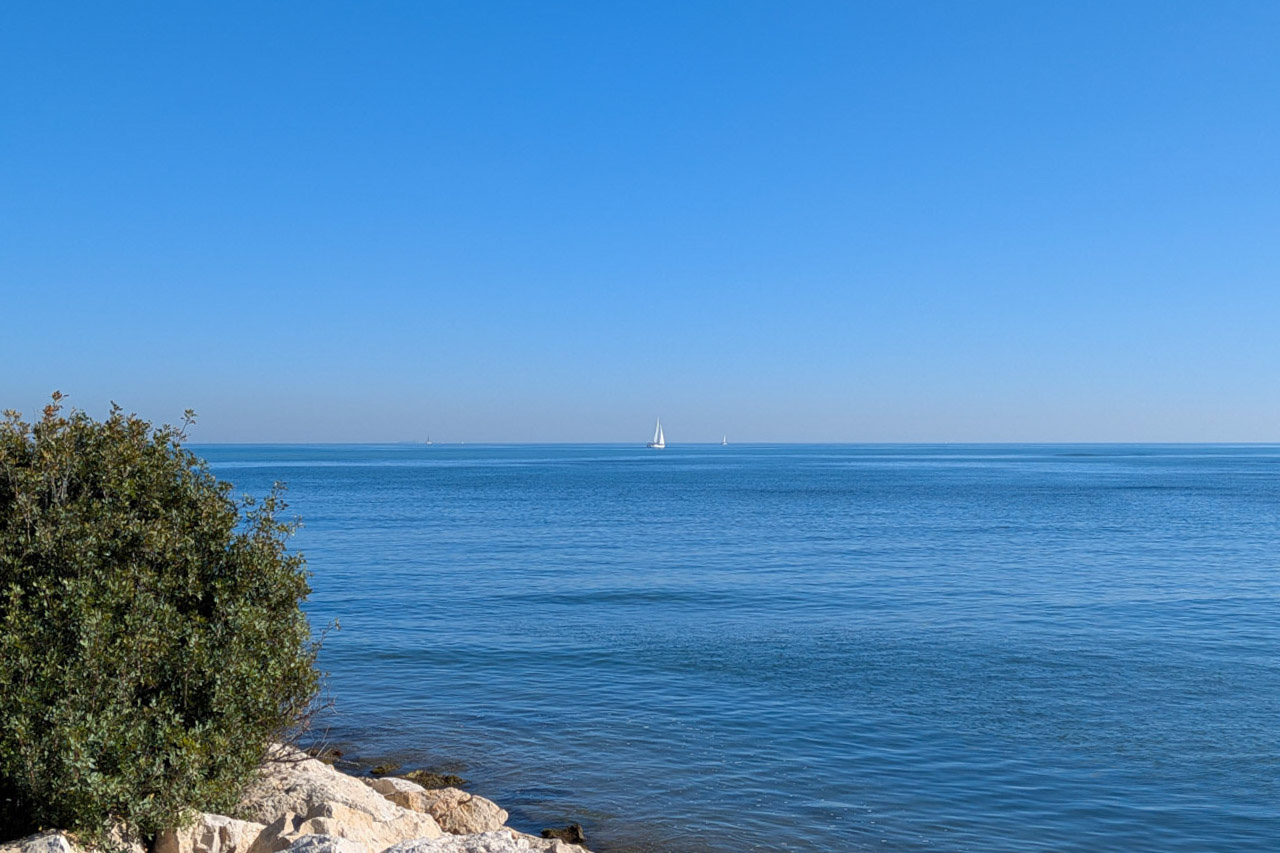 Vue panoramique sur la mer Méditerranée depuis la côte de Dénia, avec des voiliers à l'horizon et une végétation locale bordant les rochers blancs.
