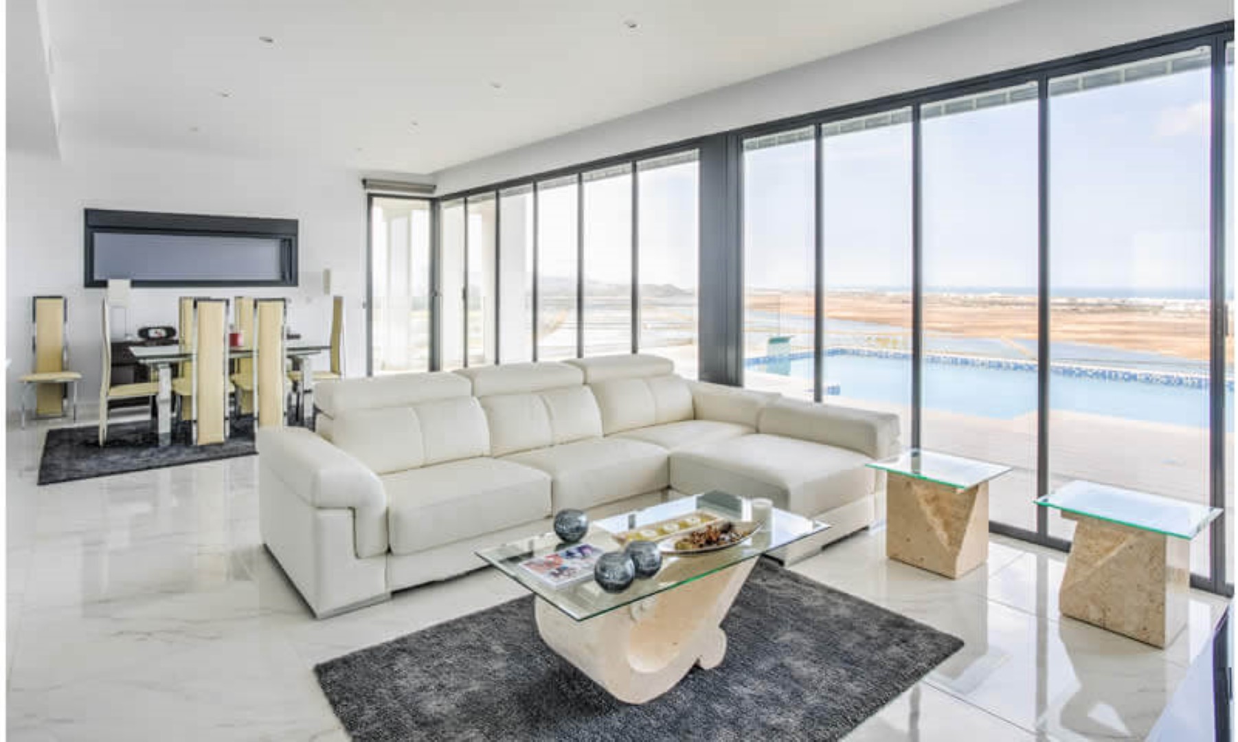 Salon-salle à manger lumineux avec sol en marbre et grandes baies vitrées offrant une vue panoramique sur la piscine à débordement et les environs.