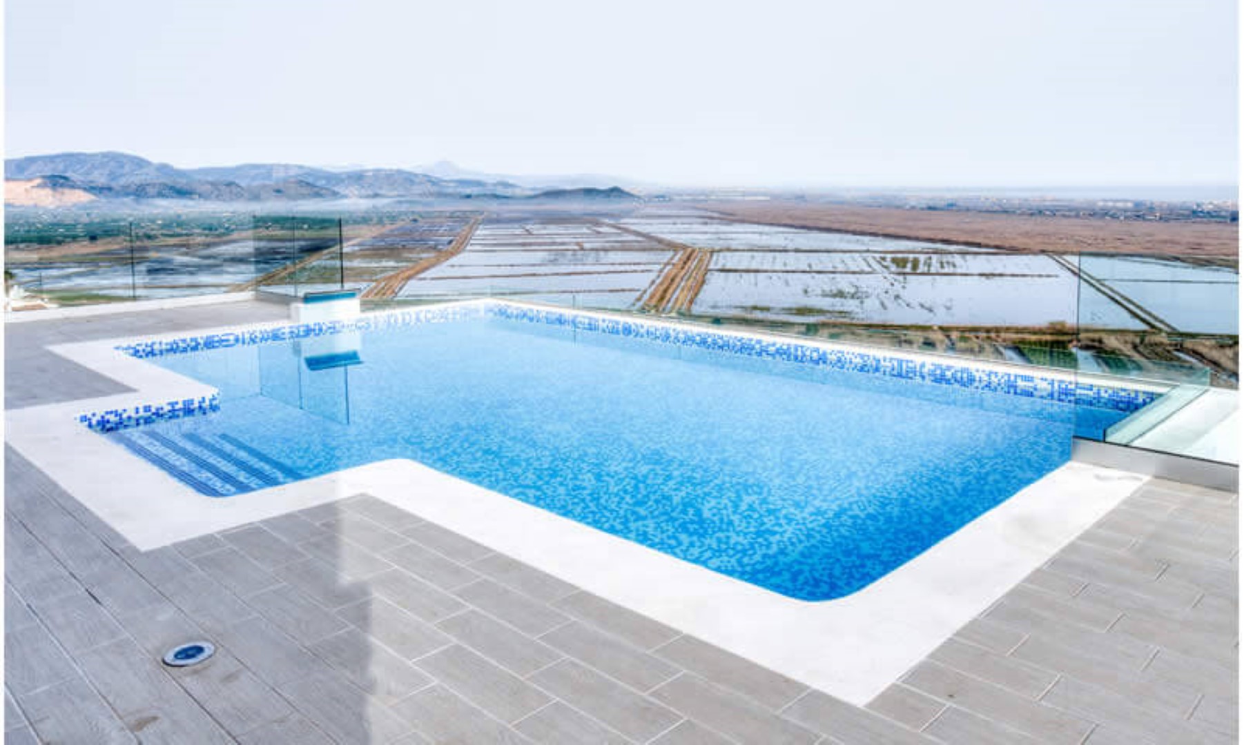 Terrasse avec piscine à débordement en mosaïque bleue et garde-corps en verre, offrant une vue panoramique sur la vallée et les montagnes environnantes.