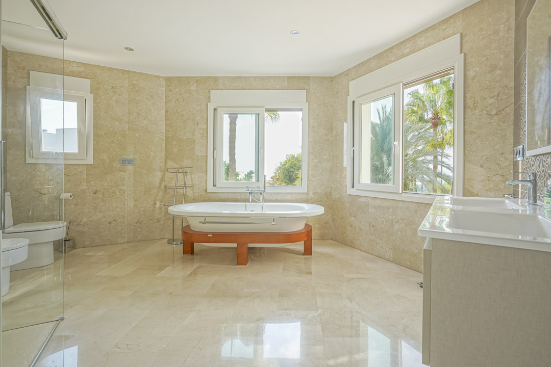 Grande salle de bain principale avec revêtement en marbre, baignoire îlot sur socle en bois et double vasque, offrant une vue sur la végétation extérieure.