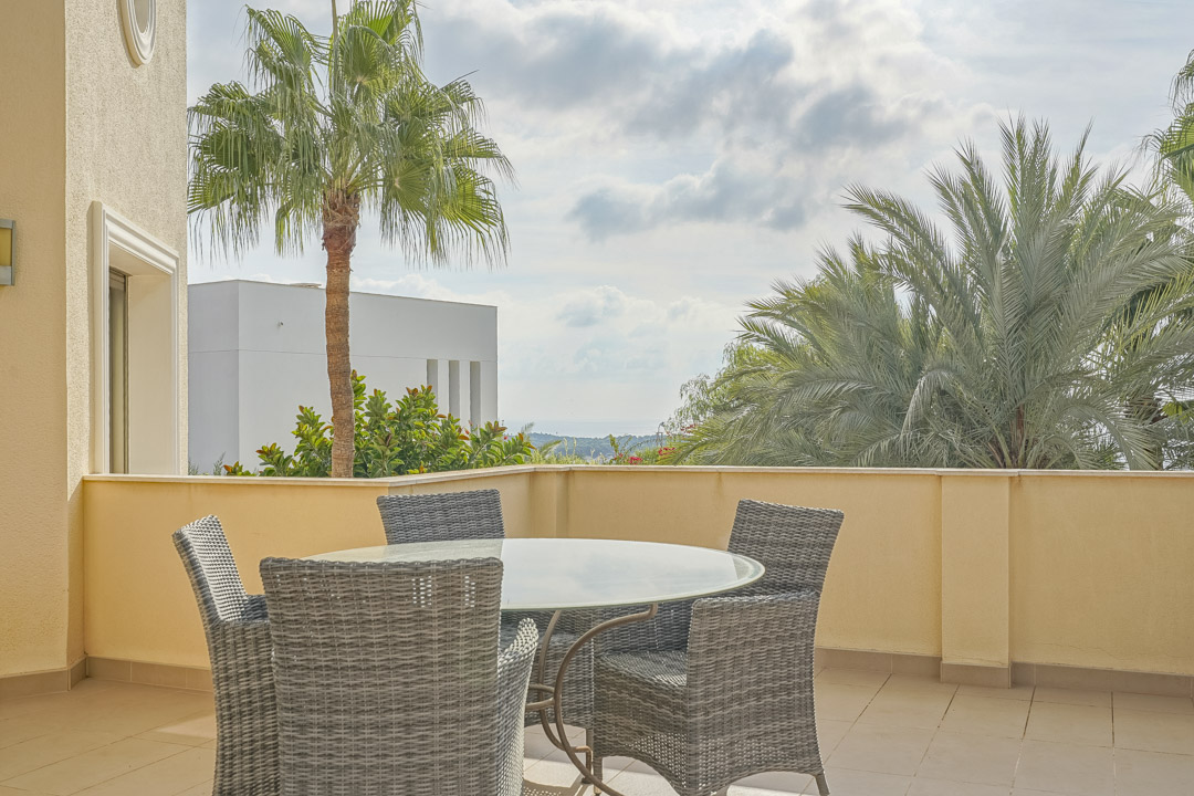 Terrasse extérieure avec table ronde en verre et chaises en osier gris, offrant une vue sur le jardin méditerranéen et la mer.