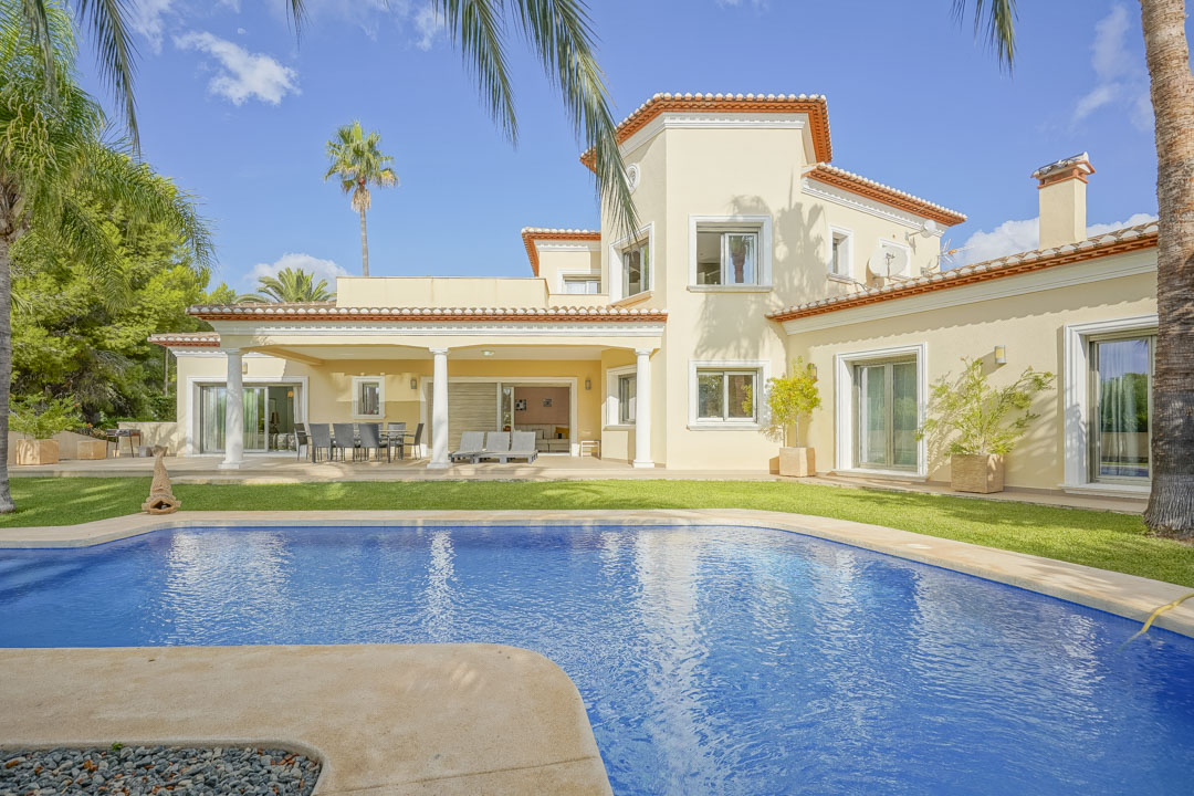Vue extérieure de la villa avec piscine, terrasse couverte soutenue par des colonnes blanches et jardin méditerranéen avec des palmiers.