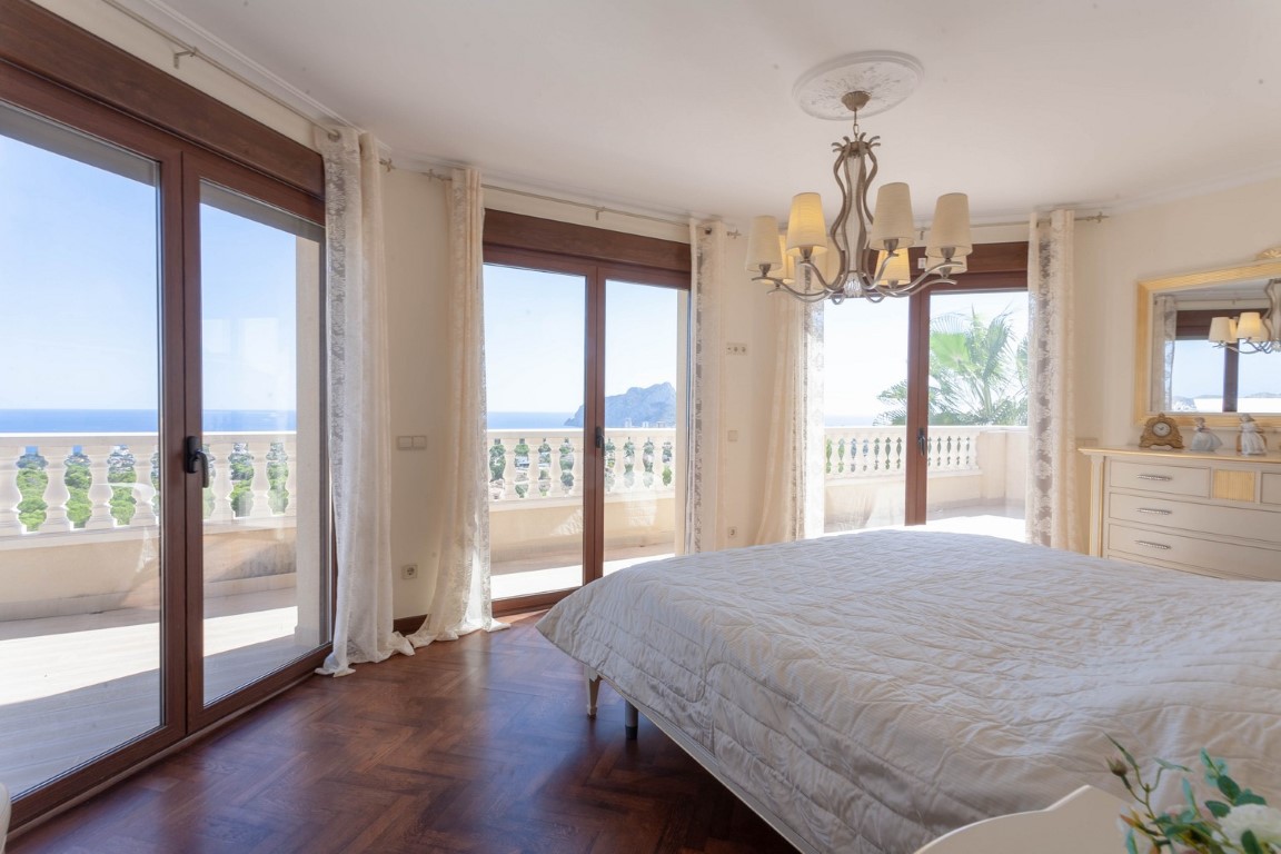 Chambre avec parquet en bois, lustre classique et grandes baies vitrées s'ouvrant sur un balcon avec vue panoramique sur la mer et le Peñón de Ifach.