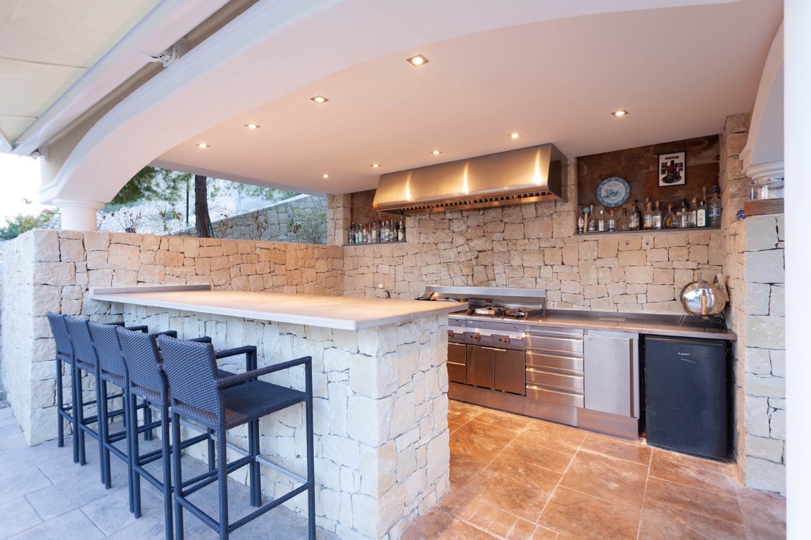 Cuisine d'été professionnelle avec murs en pierre naturelle, équipements en acier inoxydable, comptoir de bar en pierre et tabourets en rotin sous un éclairage encastré.