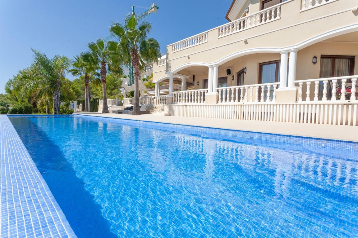 Piscine en mosaïque bleue bordant une villa méditerranéenne avec terrasses voûtées, balustrades blanches et palmiers sous un ciel clair.