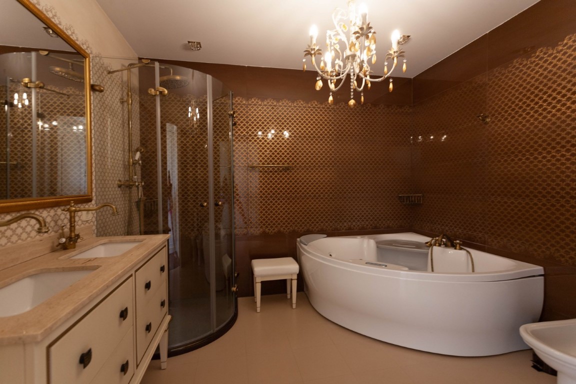 Salle de bain principale avec baignoire balnéo d'angle, douche en verre incurvée et double vasque. Les murs sont parés de carreaux marron texturés et d'un lustre classique.