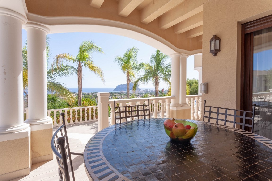 Terrasse couverte avec colonnes classiques et table en mosaïque, offrant une vue panoramique sur la mer et le Peñón de Ifach à travers les palmiers.