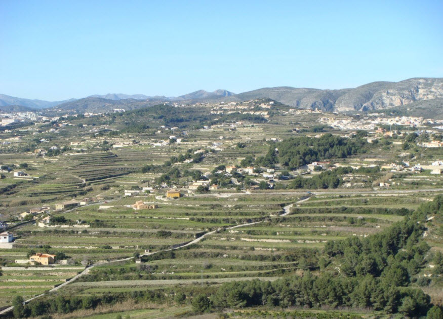 Vue panoramique de terrains en terrasses à Benitachell, mettant en valeur la topographie en paliers, la végétation méditerranéenne et les montagnes au loin.