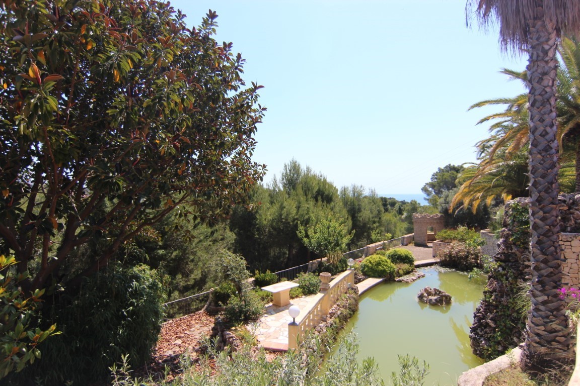 Grand bassin d'ornement avec cascade, entouré d'une végétation méditerranéenne luxuriante et de sentiers en pierre avec vue sur la mer.