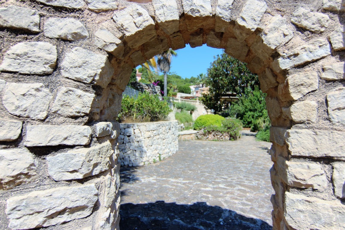 Une arche en pierre naturelle encadre l'entrée des jardins méditerranéens, avec des allées pavées et des murs de soutènement en pierre sèche.