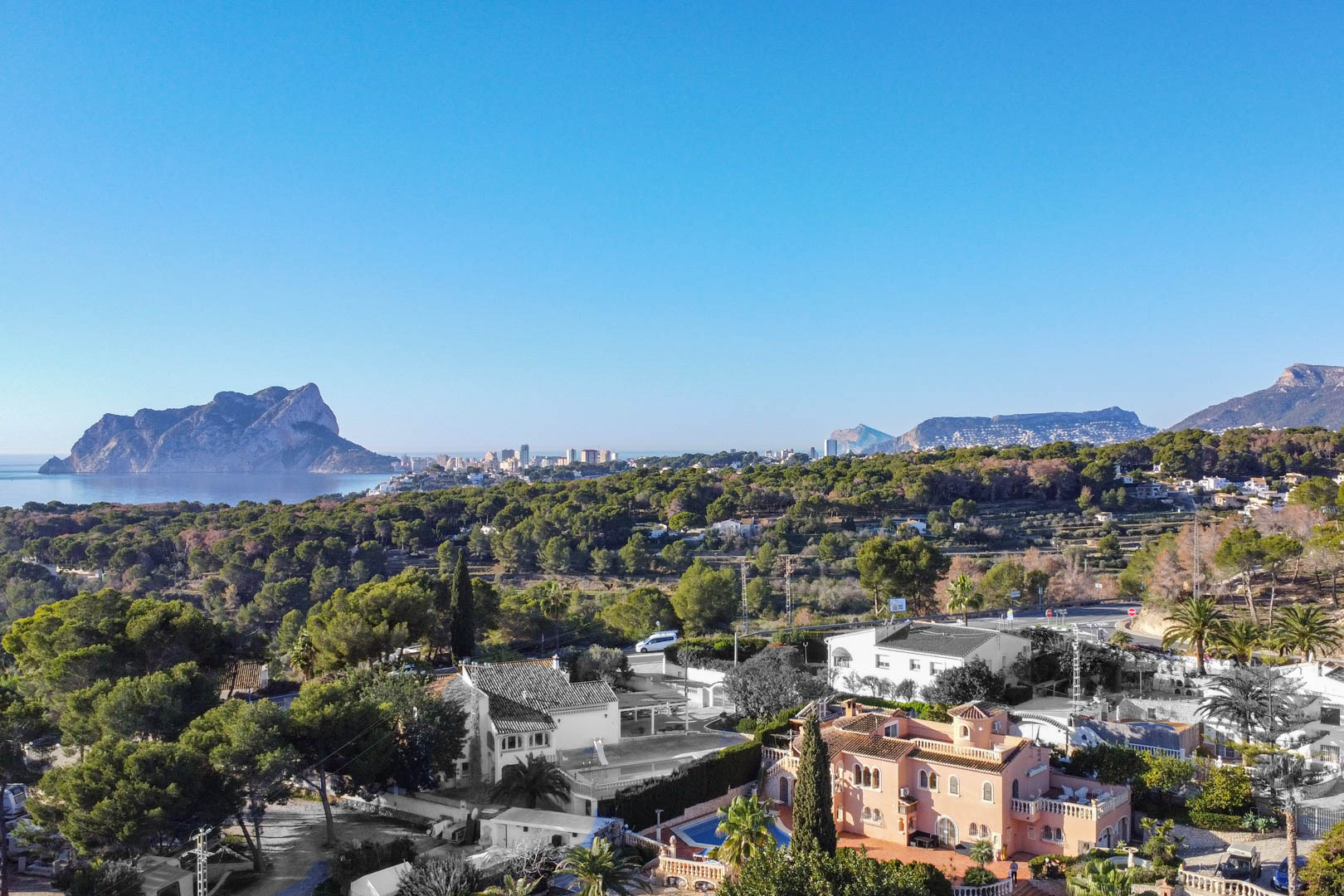 Vue panoramique aérienne sur la côte méditerranéenne avec le Peñón de Ifach, entourée d'une pinède dense et de villas résidentielles sous un ciel bleu.