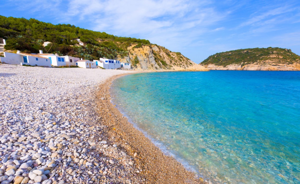 Vue panoramique de la plage de Portichol à Jávea, avec ses eaux turquoise, son rivage de galets blancs et ses maisons de pêcheurs traditionnelles au pied des falaises.