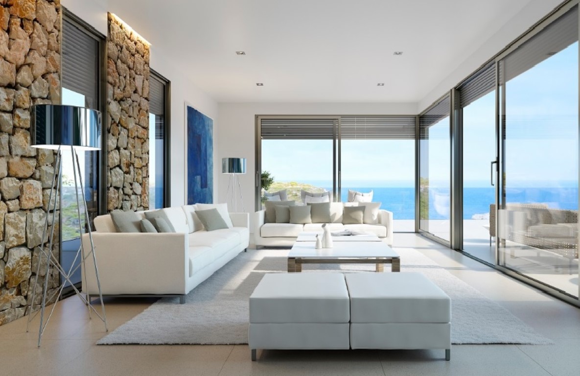 Salon lumineux avec canapés blancs, mur en pierre naturelle éclairé par LED et grandes baies vitrées offrant une vue panoramique sur la mer.