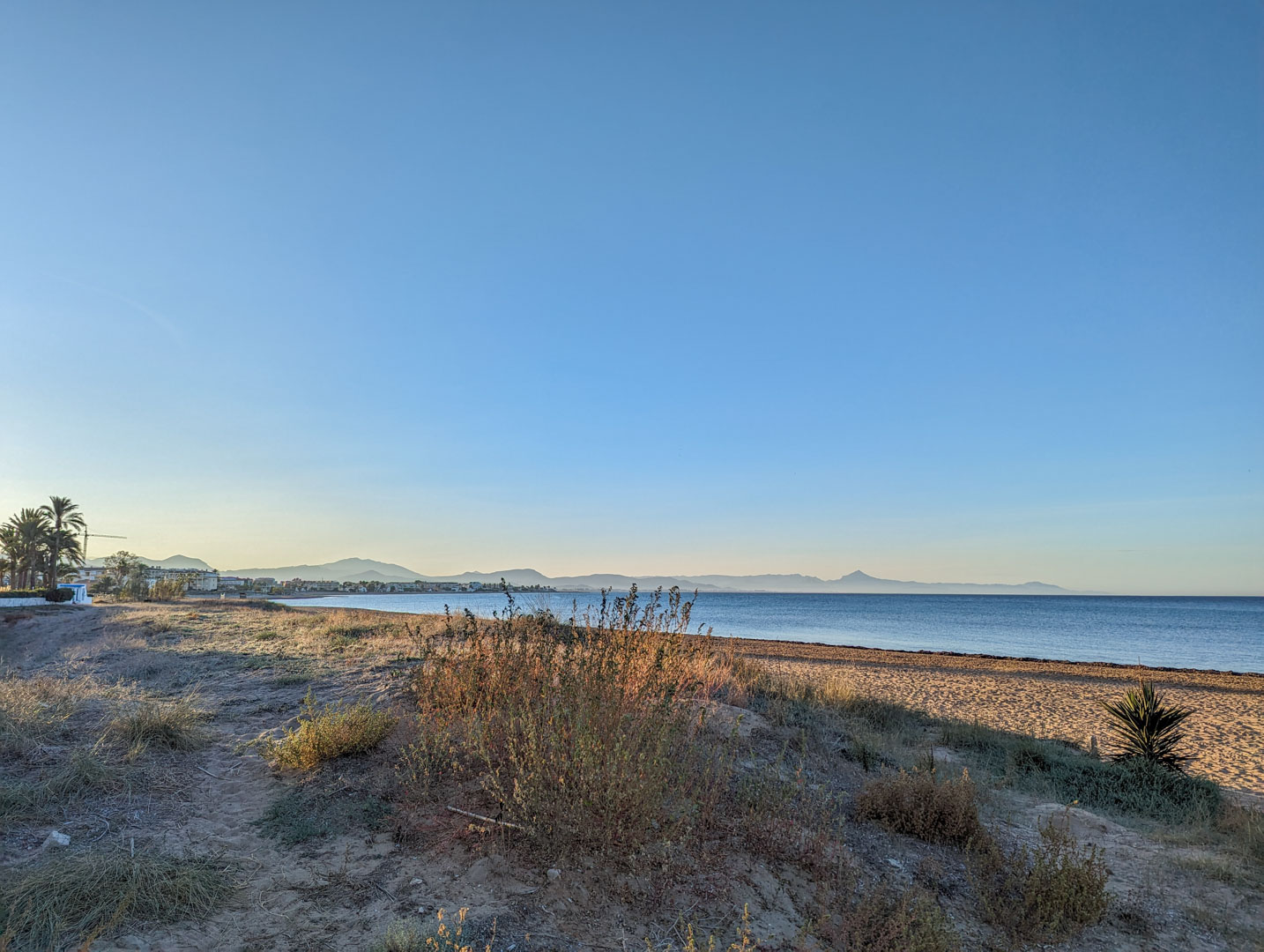 Vue de la plage naturelle à proximité, avec des dunes de sable, une végétation côtière sauvage et les montagnes en arrière-plan sous un ciel bleu.