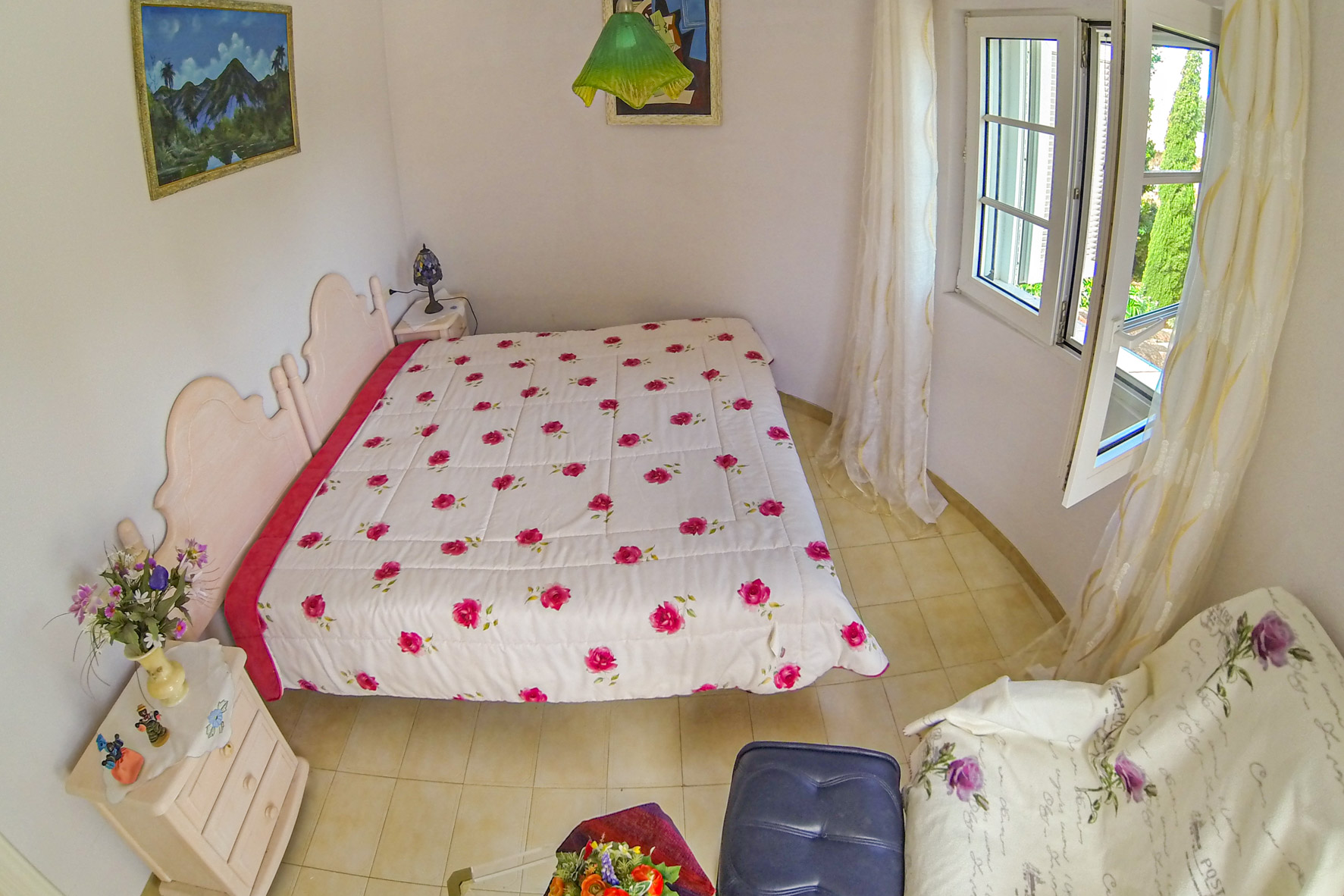 Chambre lumineuse avec lit double, literie florale et fenêtre avec rideaux transparents.