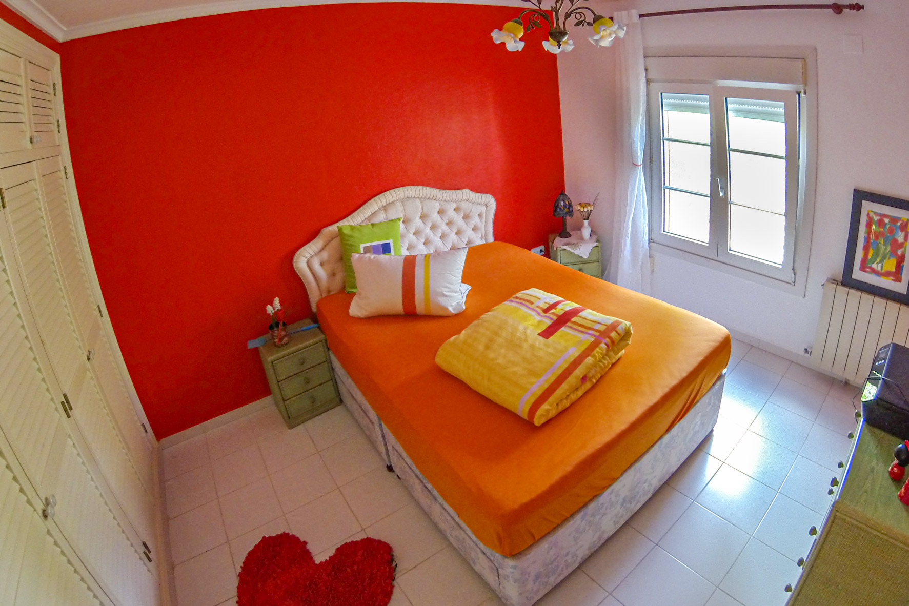 Chambre lumineuse avec mur rouge, lit double avec linge orange et tête de lit rembourrée.