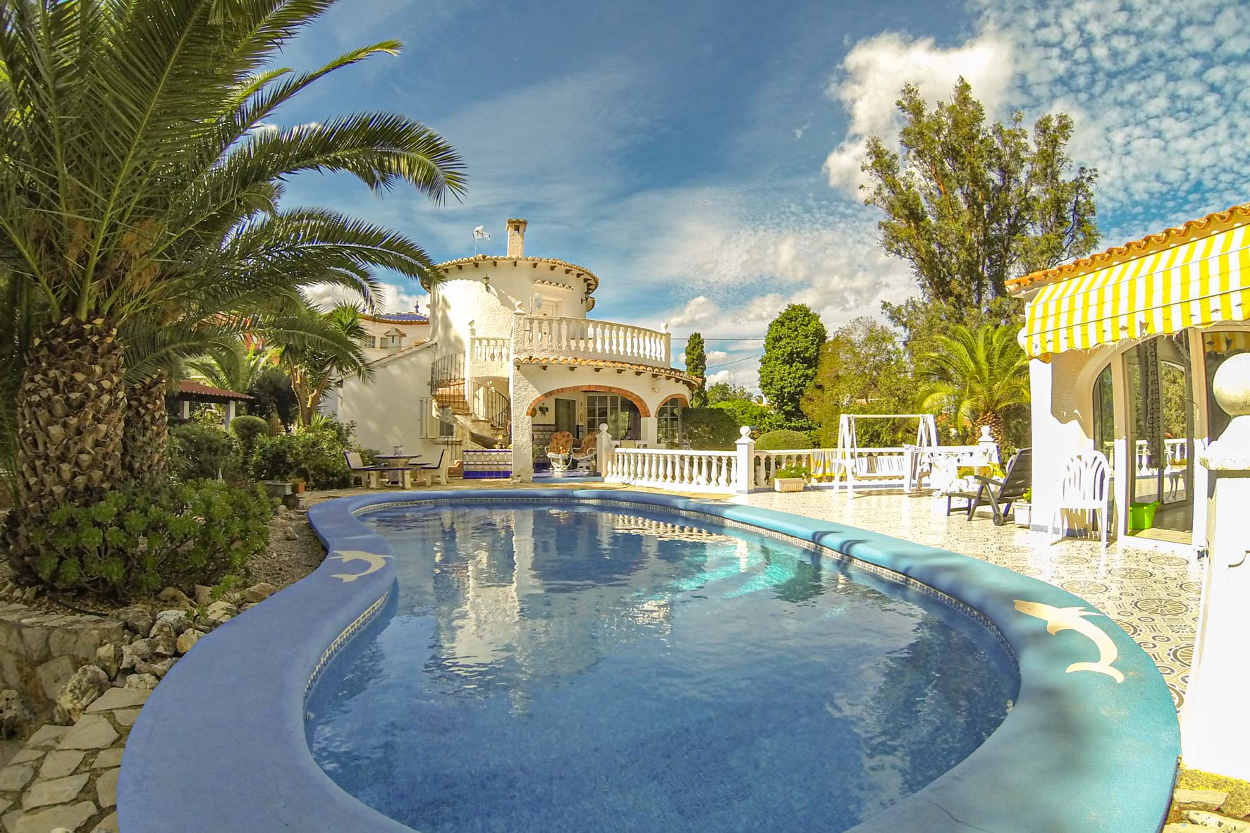 Vue de la piscine bleu turquoise avec bordure décorée de dauphins et de la façade de la villa blanche à deux étages.
