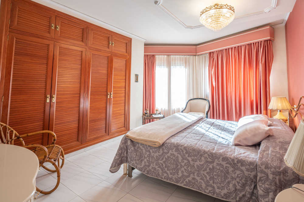Chambre à coucher avec placards intégrés en bois, grande fenêtre aux rideaux rosés, sol en carrelage clair et lustre en cristal raffiné.