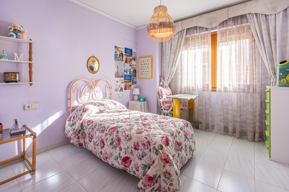 Chambre individuelle aux murs lavande et sol en carrelage clair. Elle dispose d'un lit avec tête de lit en fer forgé et d'un bureau près de la fenêtre en bois.