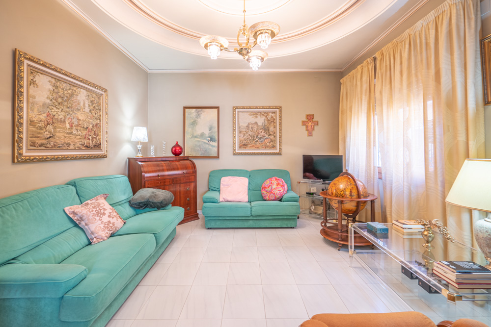 Salon lumineux avec canapés turquoise, carrelage blanc et moulures circulaires au plafond. Décoré avec des meubles en bois classiques, un secrétaire et des tapisseries encadrées.