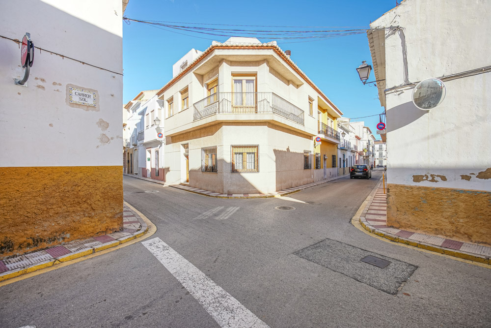 Vue extérieure d'angle d'une maison de village traditionnelle à deux étages, avec balcon en fer forgé et fenêtres sécurisées dans une rue calme et ensoleillée.