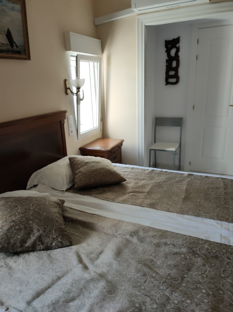 Chambre avec lit double, tête de lit en bois classique et climatisation. Elle offre une lumière naturelle, une table de chevet et un coin avec une chaise d'appoint près de la porte.