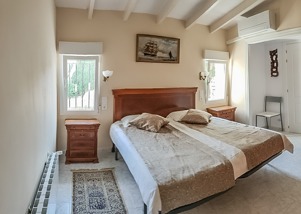 Chambre avec plafond à poutres apparentes blanches, lit double en bois, tables de chevet assorties, climatisation et chauffage par radiateur.