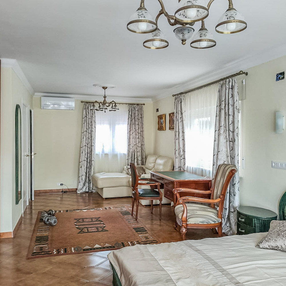 Chambre spacieuse avec coin salon, bureau en bois classique, climatisation et sol en grès. Les grandes fenêtres avec rideaux à motifs offrent une luminosité naturelle.