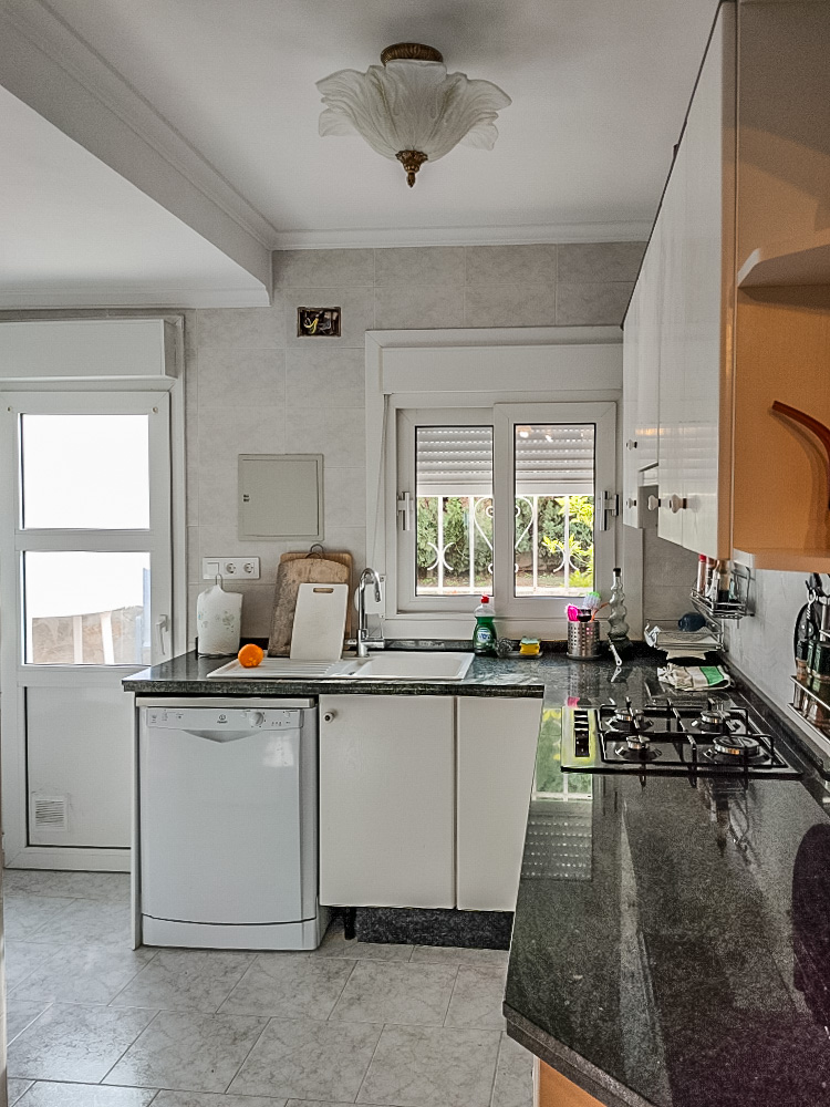 Cuisine pratique avec plan de travail en granit noir, plaque de cuisson au gaz et meubles blancs. Elle offre un accès direct à l'extérieur et une lumière naturelle.