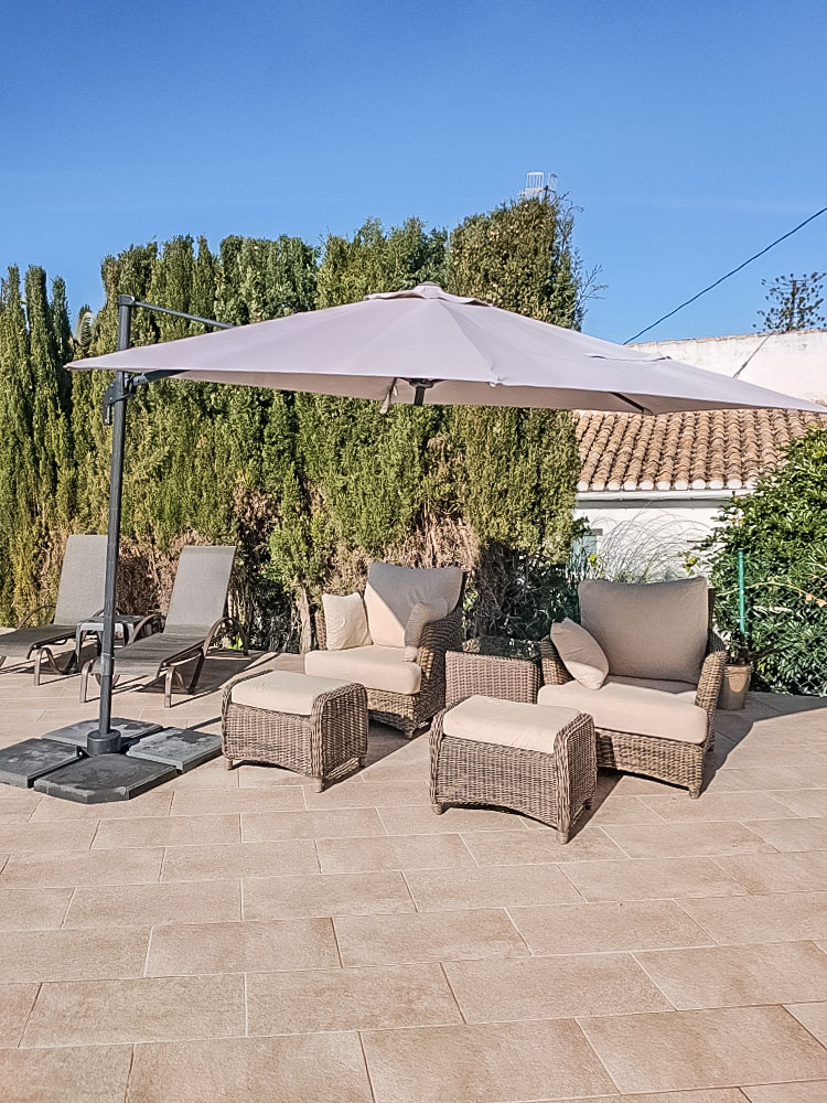 Espace de détente extérieur avec fauteuils en résine tressée, repose-pieds et grand parasol déporté sur une terrasse pavée bordée de hautes haies.