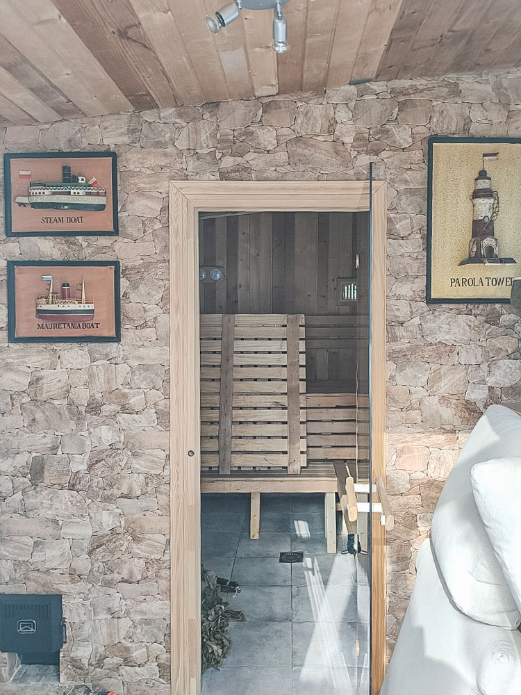 Sauna privé avec murs en pierre naturelle et porte vitrée. L'intérieur est entièrement revêtu de bois avec des bancs à plusieurs niveaux et des instruments de mesure.