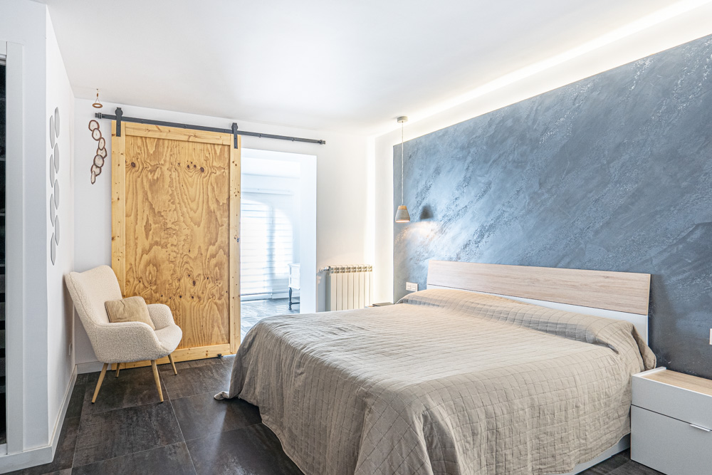 Chambre avec mur d'accent gris texturé, éclairage LED indirect, sol en carrelage foncé et porte coulissante en bois naturel sur rail métallique noir.